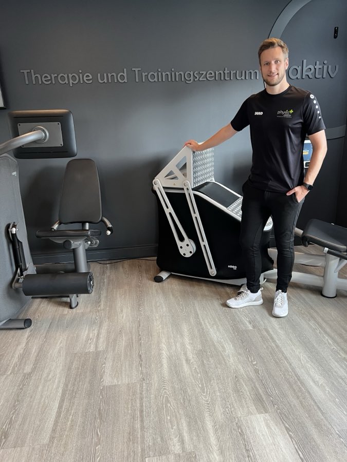 Physiotherapeut im Praxis-Shirt im Trainingsbereich vor dem Schriftzug Therapie und Trainingszentrum aktiv